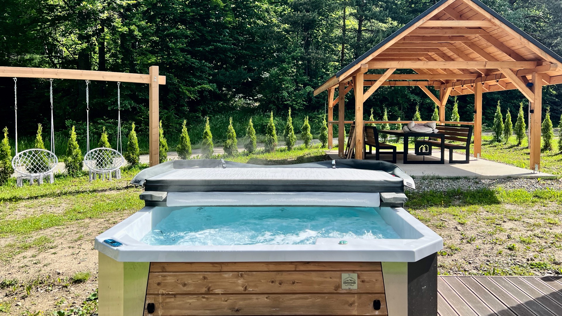 Otwarte jacuzzi z wodą latem, za nim altana ze stołem i huśtawki na tle lasu