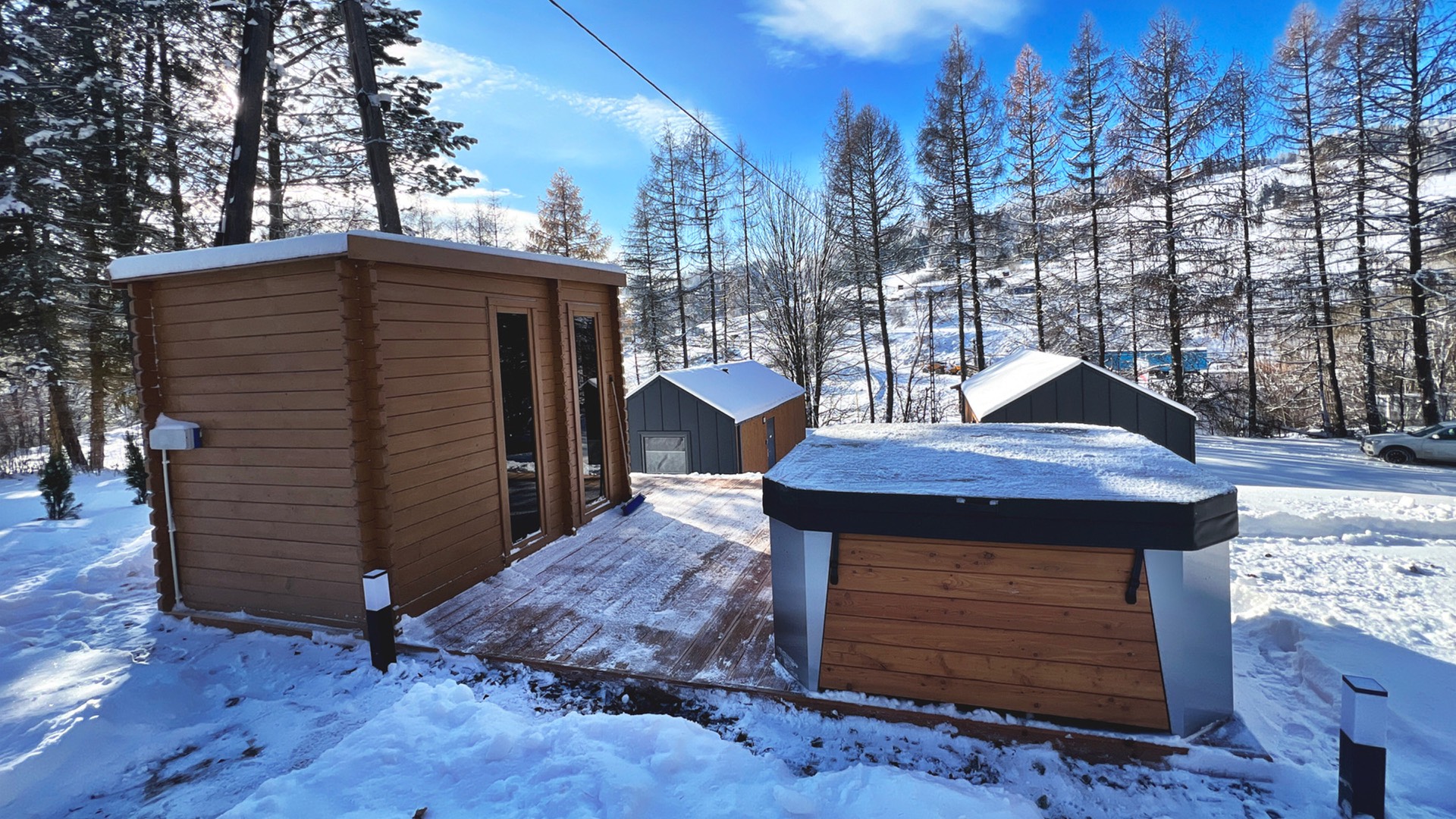 Sauna i jacuzzi na drewnianym podeście w śniegu, w tle domki i ośnieżony stok